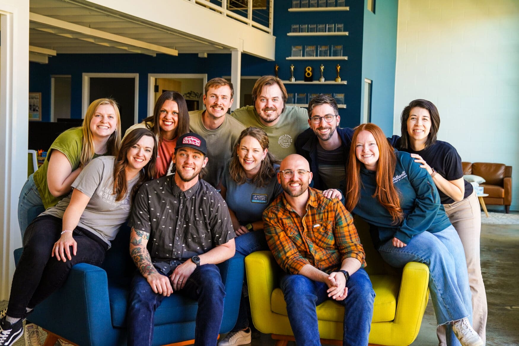 A large group of eleven coworkers are gathered closely together, smiling for a team photo in a modern office space. Two men sit in blue and bright yellow armchairs in the foreground, while the rest of the team leans in behind and around them. A deep blue accent wall with shelves displaying awards is visible in the background.
